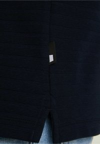 Dunkelblaues strukturiertes Gewebe mit einem seitlichen Schlitz und einem schwarzen Etikett mit einem weißen Quadrat am Saum.