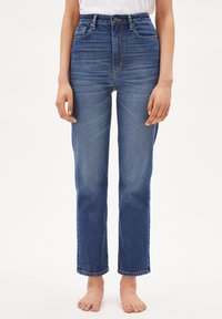 Kvinna i högmidjade blå denimjeans och en vit topp, stående barfota mot en enfärgad vit bakgrund.