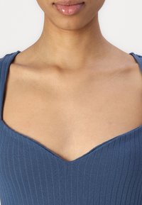 Modrý žebrovaný top s výstřihem do V, který se vyznačuje hladkou texturou a přiléhavým střihem. Materiál se zdá být elastický a tvarující.
