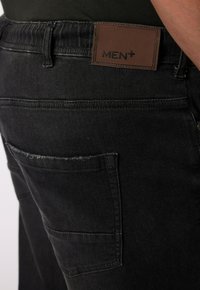 Vaqueros de mezclilla negra con una superficie texturada, que presentan un parche de cuero marrón etiquetado como "MEN+" y un bolsillo trasero con ligero desgarro.