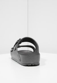 Svart gummisandal med öppen bak, strukturerad sula och överdragrem med en rosettaccent. Slät yta och flexibel design.