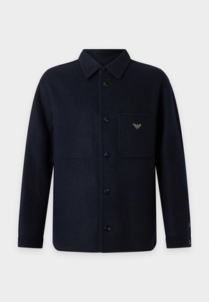 Homme portant une veste noire boutonnée par-dessus un col roulé gris, un jean bleu et des chaussures noires, debout les mains dans les poches sur fond blanc.