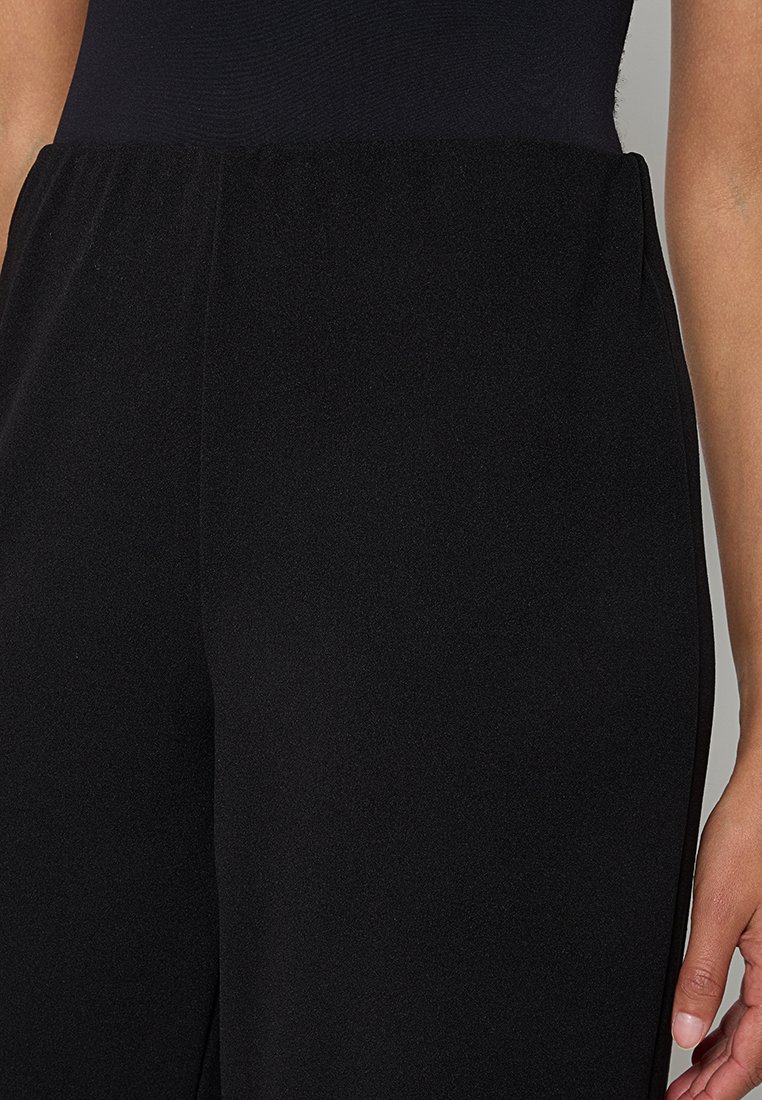 Gros plan sur une personne portant un pantalon noir taille haute avec une ceinture élastique, vu des hanches au milieu de la cuisse sur fond neutre.