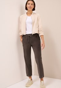 Veste bomber beige clair, t-shirt blanc, pantalon à cordon gris foncé et baskets beiges. Texture lisse, coupe décontractée et design minimaliste.