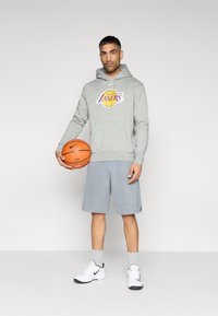Sweat à capuche gris avec logo des Lakers, associé à un short de sport gris clair. Tenant un ballon de basket orange, portant des baskets blanches avec des accents noirs.
