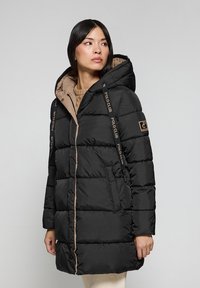 Svart puffjacka med en beige foder, med huva, horisontellt kviltad design, logotypdetalj på axeln och dragkedjefickor.