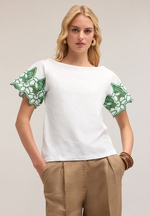 T-shirt bianca in cotone con bordo in pizzo verde sulle maniche. Vestibilità morbida, scollatura rotonda, abbinata a pantaloni color sabbia.