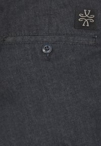 Tessuto denim blu scuro con una sottile texture, dotato di chiusura con bottone sulla parte posteriore e un'etichetta con logo ricamato nell'angolo in alto a destra.