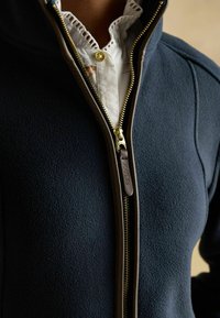 Námornícky fleece kabát s hnedým koženým zipsom čiastočne otvoreným, ukazuje biele tričko s čipkovým golierom a zlatým gombíkom pod ním.