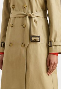 Beigi trenčkot ar jostu, divi priekšējo pogu slēgšanas dizains, lielas dzeltenas pogas un atloka aizdare, ar gludu tekstūru un pielāgotu piegriezumu.