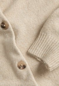 Cardigan in lana beige con due bottoni marroni marmorizzati e una manica con polsino a coste, che mostra la texture del tessuto e i dettagli delle cuciture.