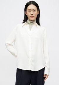 Joven mujer con cabello negro liso, vistiendo una camisa blanca de botones sobre un suéter de cuello alto a rayas y pantalones negros, de pie con la mano en la cadera.