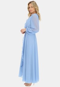 Jasnoniebieska długa sukienka z falbaniastym przodem, długimi przezroczystymi rękawami i zwiewnym materiałem. Połączona z jasnobeżowymi sandałkami na szpilkach.