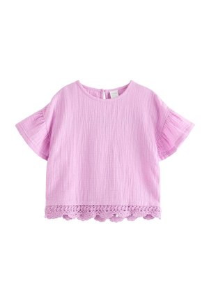 Smilende ung pige med blondt hår iført en lyserød top med flæser og korte ærmer samt matchende bukser med vide ben og blondedetaljer, stående indendørs.