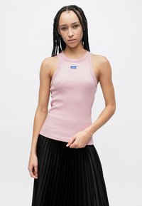 Világos rózsaszín bordás anyagú ujjatlan felső kerek nyakkivágással és kék logóval, fekete pliszírozott szoknyával párosítva. Egyszerű és testhez simuló szabás.