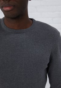 Szary sweter dzianinowy z okrągłym dekoltem, o gładkiej teksturze i dopasowanym kroju, ukazany na tle jasnej cegły.