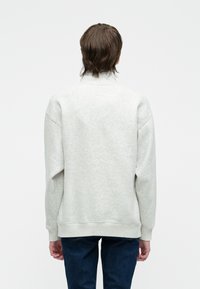 Osoba o średniej długości brązowych włosach, ubrana w jasnoszary sweatshirt i ciemnoniebieskie dżinsy, widziana od tyłu na białym tle.