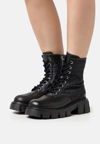 Des bottes de combat noires présentent un dessus en cuir verni texturé avec des lacets, une fermeture éclair latérale et une semelle en caoutchouc épaisse. Motif de logo notable sur les côtés.