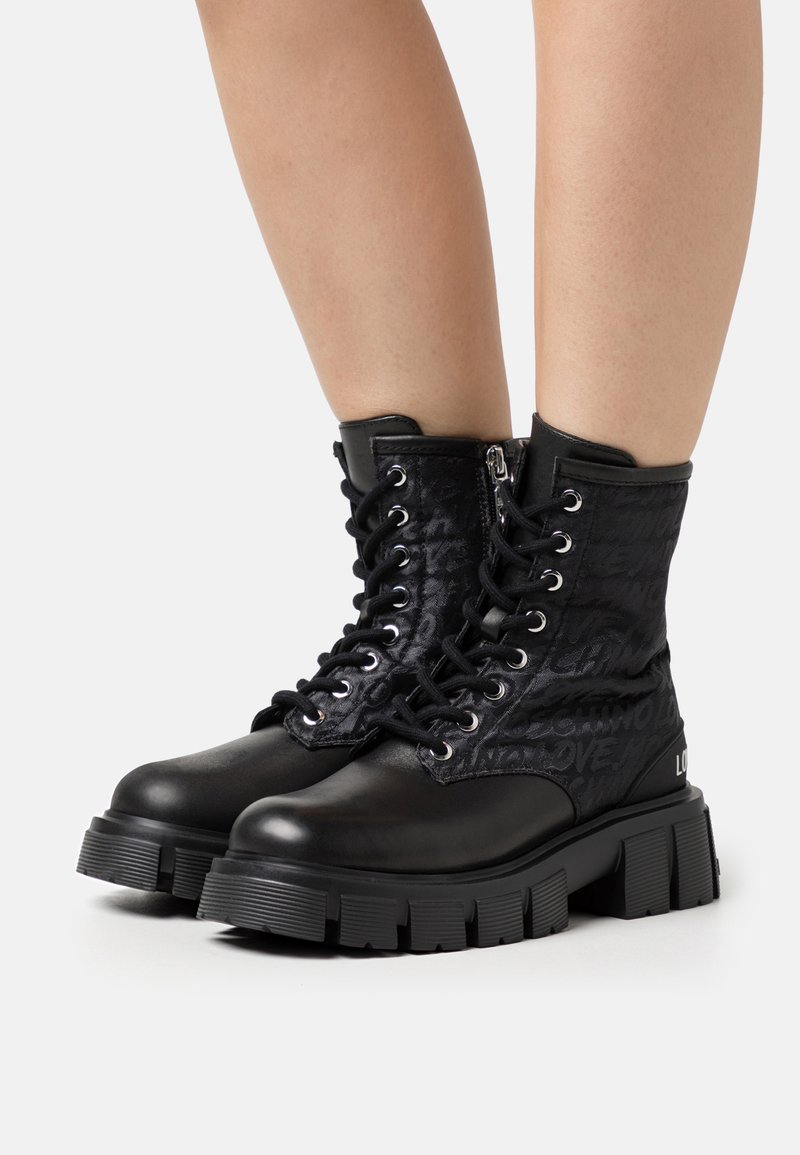 Des bottes de combat noires présentent un dessus en cuir verni texturé avec des lacets, une fermeture éclair latérale et une semelle en caoutchouc épaisse. Motif de logo notable sur les côtés.