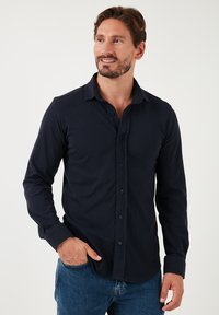 Cămășă albastră închis cu mânecă lungă, dintr-un material moale, având guler standard, o singură linie de nasturi și o croială lejeră.