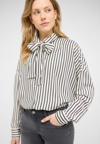 Striped blouse in white and navy, featuring a bow tie collar, button closure, and long sleeves with cuff detailing. Paired with dark jeans.