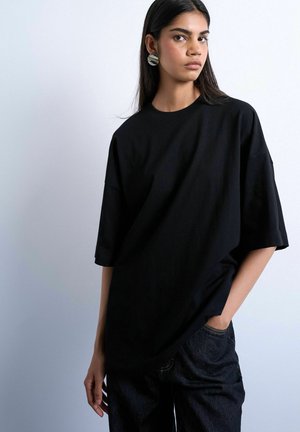 Young woman with long dark hair wearing large round earrings, an oversized black T-shirt, and dark jeans, posed against a plain light background.