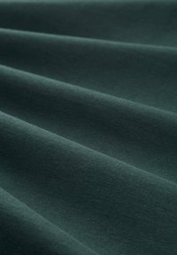 Ciemnozielony materiał o gładkiej, matowej teksturze, z lekkim drapowaniem i subtelną głębią od fałd, ukazujący swoje miękkie właściwości.