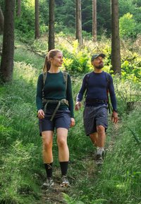Twee wandelaars op een bospad. De een draagt een groen shirt met lange mouwen en zwarte korte broek; de ander een blauw shirt met lange mouwen en grijze korte broek.