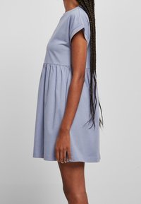 Frau mit langen, geflochtenen Haaren, die ein kurzärmeliges, lockeres, hellblaues Kleid trägt und vor einem schlichten hellen Hintergrund steht.