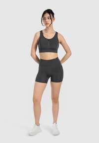 Zwarte sportbra met een rits aan de voorkant en bijpassende high-waisted shorts, gemaakt van rekbaar materiaal. Model staat met handen op de heupen tegen een effen achtergrond.