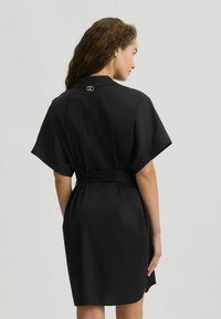 Abito nero con vita in vita, maniche corte, colletto pieghettato e un accento dorato vicino al collo. Tessuto liscio con un design minimalista.