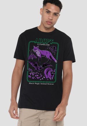 Junger Mann trägt ein schwarzes T-Shirt mit lila Illustrationen eines Fuchses, einer Fledermaus, einer Schlange, eines Waschbären, einer Spinne und eines Nachtfalters mit dem Text "Adopt a Familiar".