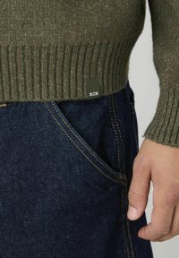 Pull en maille vert olive avec finition côtelée, étiquette montrant "BZB". Porté avec un jean en denim bleu foncé, mettant en avant les coutures des poches et la texture.