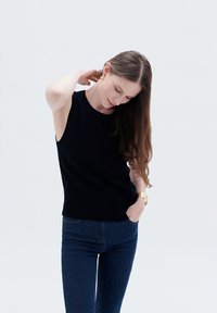 Top senza maniche nero con scollo rotondo, abbinato a jeans blu scuro. La modella ha i capelli lunghi e si sta aggiustando i capelli.