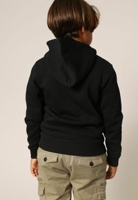 Zwart capuchontrui met een losse pasvorm en geribbelde zoom, gedragen over kaki cargobroek met knoopaccenten. De stof lijkt zacht en comfortabel.