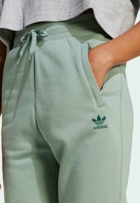 Green cotton joggers with ribbed waistband and tie adjustment. Features side pockets and a dark green embroidered logo on the leg.