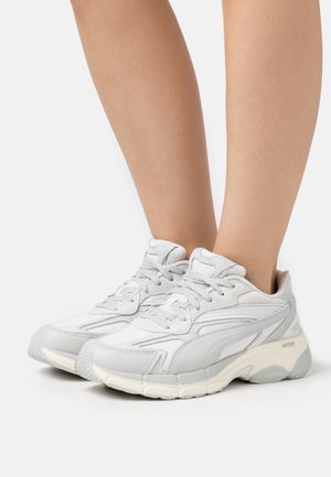 Zapatillas deportivas blancas que combinan materiales de cuero y malla, diseño redondeado, acentos grises, suela texturizada y cordones.