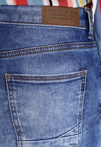 Takapocket sinisistä Esprit-farkuista, joissa on näkyvät saumat, ja värikkään raidallisen paidan helma on sisätetty.