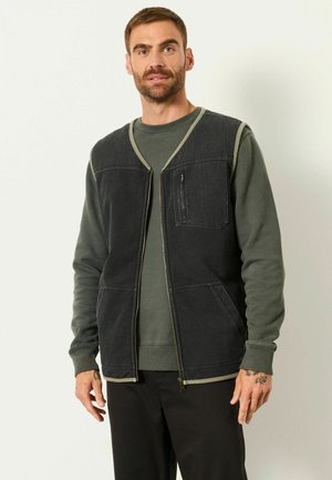 Mann mit kurzen Haaren und Bart, trägt eine dunkle Reißverschlussweste über einem grünen Langarmshirt und schwarze Hose, steht vor einem einfarbigen Hintergrund.