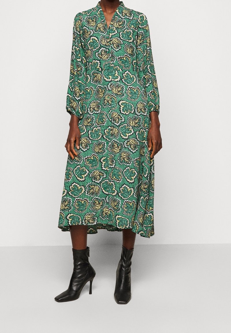 Personne portant une robe midi verte avec des motifs de feuilles jaunes et noires et des bottines noires à talons aiguilles.