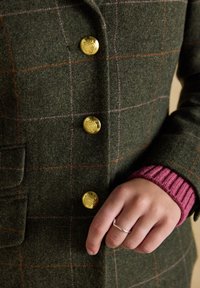 Hand met zilveren ring op een donkergroene geruite jas met gouden knopen en een bordeauxrode geribde trui-mouw zichtbaar bij de pols.