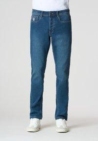 Jeans in denim blu con una vestibilità dritta, dotati di cinque tasche, una leggera scoloritura e una patch del marchio sulla tasca posteriore. Indossati con sneakers bianche.