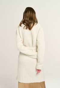 Strickjacke in Creme mit taillierter Silhouette, langen Ärmeln und geripptem Saum. Der Stoff wirkt weich und strukturiert. Rückansicht, getragen über einem beigen Kleid.