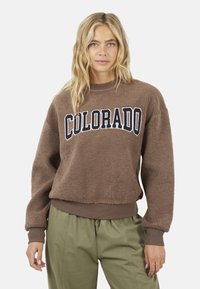 Donna bionda che indossa una felpa in pile marrone con la scritta "COLORADO" e pantaloni verde oliva, in piedi su uno sfondo neutro.