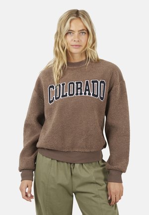Donna bionda che indossa una felpa in pile marrone con la scritta "COLORADO" e pantaloni verde oliva, in piedi su uno sfondo neutro.