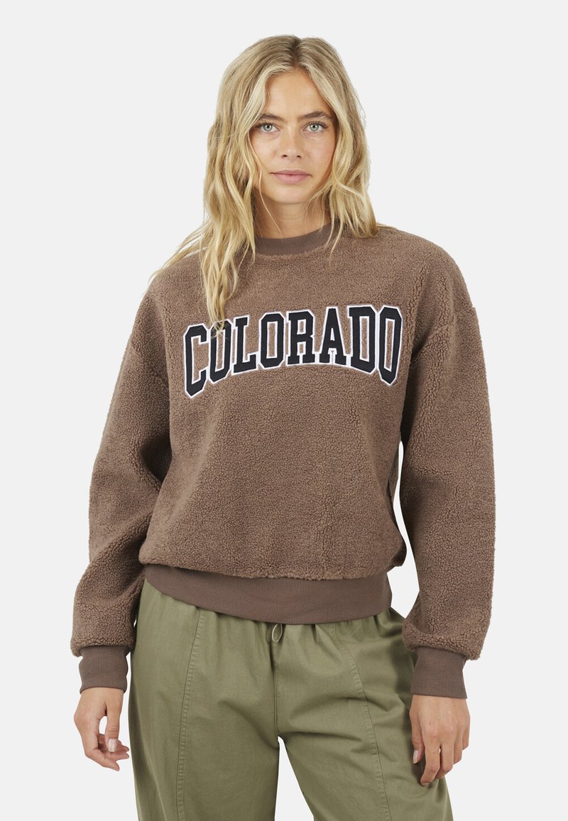 Donna bionda che indossa una felpa in pile marrone con la scritta "COLORADO" e pantaloni verde oliva, in piedi su uno sfondo neutro.