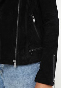 Czarna zamszowa kurtka z poziomymi przeszyciami, dwoma bocznymi kieszeniami na zamek oraz srebrnymi zamkami błyskawicznymi z przodu i na rękawach.