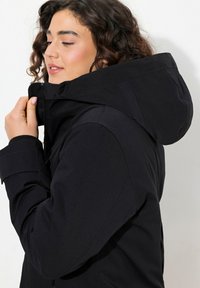Svart huva-jacka med en slät, matt yta som har elastiska ärmslut och zip-försedda frampocketar. Designad för värme och funktionalitet.