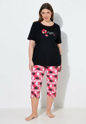 Nainen seisoo paljain jaloin, yllään musta lyhythihainen paita, jossa on "dream on" -teksti, ja vaaleanpunaiset kukkakuosilliset caprihousut.