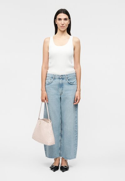 Γυναίκα που φοράει λευκή αμάνικη μπλούζα, φαρδιά ανοιχτό μπλε τζιν, μαύρα μυτερά μπαλαρίνες, κρατώντας μία ανοιχτό ροζ πλεκτή τσάντα πάνω σε λευκό φόντο.